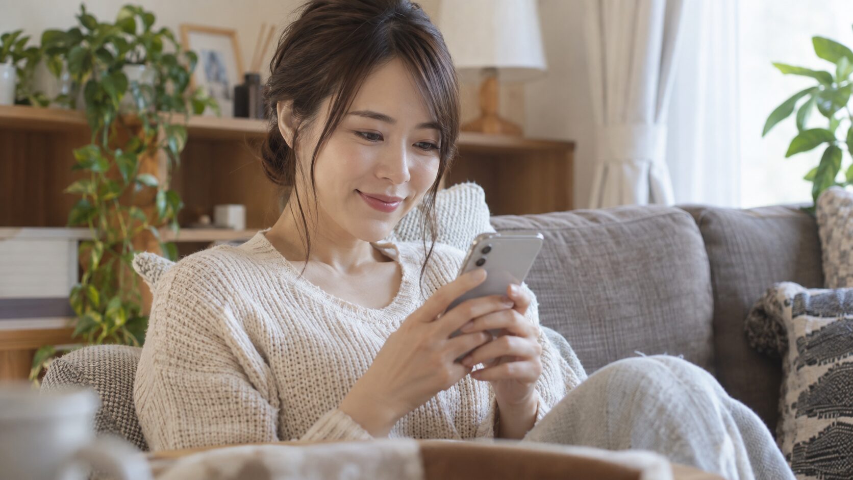 自宅のソファで笑顔でスマートフォンを操作する女性
